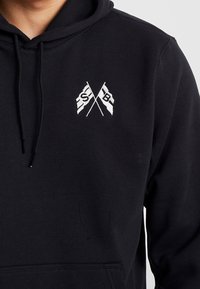 Svart hoodie med en framfickan, prydd med vita broderade korsade flaggor och bokstäverna "S" och "B." Mjuk tyg, justerbar dragskohuva.