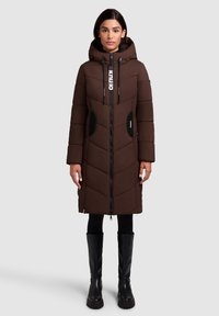 Caban puf maro cu glugă, având un sistem de închidere cu fermoar, buzunare negre ca accent și un model în zigzag. Asortat cu cizme negre până la genunchi.