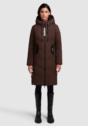 Brauner Puffer-Mantel mit Kapuze, mit einem Reißverschluss, schwarzen Akzenttaschen und einem Zickzackmuster. Kombiniert mit schwarzen kniehohen Stiefeln.