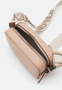 Ružová kožená crossbody taška so zipsom, zdobená polkruhovým perforovaným dizajnom a nastaviteľným plátenným popruhom s logom.