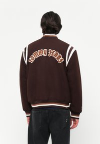 Persona con capelli scuri che indossa una giacca varsity marrone con strisce bianche e il testo "Tommy Jeans" sulla schiena, abbinata a jeans neri.