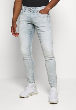 Mann trägt hellblaue Skinny-Jeans, schwarzes Hemd, weiße Socken und weiße Turnschuhe, steht mit einer Hand in der Tasche.