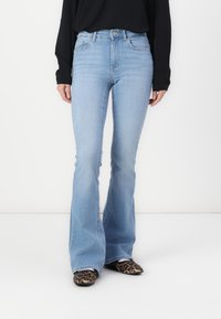 Femme portant un jean évasé taille haute bleu clair, un haut noir à manches longues et des chaussures à bout pointu imprimé léopard, debout devant un fond blanc.