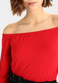 Rode off-shoulder top van zachte stof met een aansluitend ontwerp en een gerimpelde halslijn. Gecombineerd met een zwarte tailleband die aan de onderkant zichtbaar is.