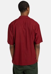 Camicia a maniche corte color bordeaux realizzata in tessuto morbido; presenta un colletto basso e una vestibilità comoda, mostrata dal retro.