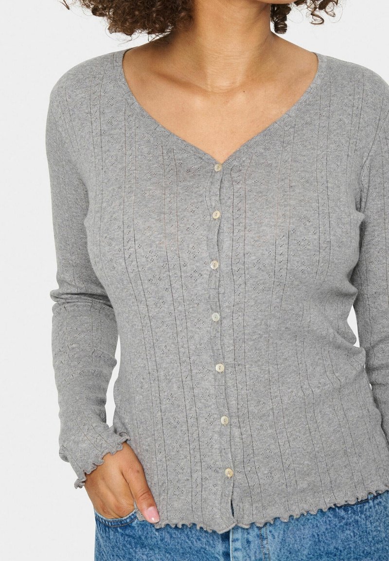 Femme portant un cardigan gris clair boutonné avec des motifs verticaux texturés et des bords volantés, associé à un jean en denim bleu.