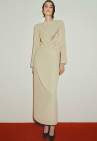 Beige jurk met lange mouwen, voorzien van een cascade van franje aan één kant en een strakke, aansluitende pasvorm. Gekocht in combinatie met zwarte, puntige hakken.