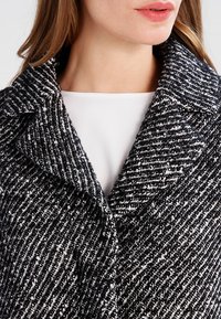 Cappotto cesellato in bianco e nero con motivo a spina di pesce, dotato di colletto a punta e chiusura con bottoni, posizionato su una maglietta bianca.