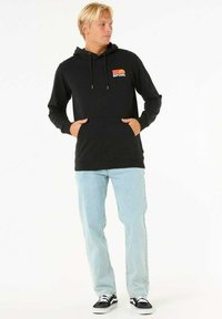 Zwarte hoodie met een voorzak en een koord in de capuchon. Voorzien van een meerkleurig "RIP CURL" logo op de borst. Gecombineerd met lichtblauwe jeans.