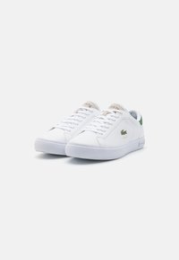 Bílé tenisky s hladkým koženým svrškem, zelenými akcenty na patě a texturovanou gumovou podrážkou. Logo Lacoste na boku.