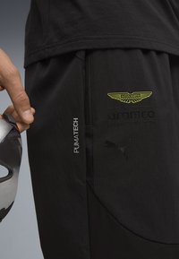 Schwarze Sporthosen aus strukturiertem Material, mit Aston Martin-Logo, PumaTech-Branding und einem eleganten Taschen-Design. Silberne Akzente an der Tasche.