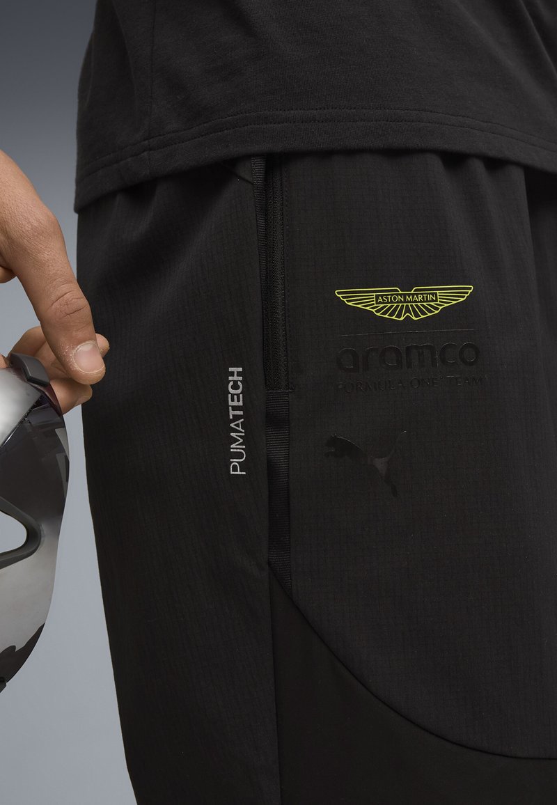 Schwarze Sporthosen aus strukturiertem Material, mit Aston Martin-Logo, PumaTech-Branding und einem eleganten Taschen-Design. Silberne Akzente an der Tasche.