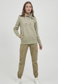 Felpa verde oliva con cappuccio e coulisse, tasca a marsupio, abbinata a pantaloni cargo beige con polsi elasticizzati e tasche laterali. Sneakers bianche.