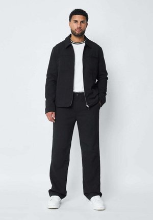 Veste noire zippée sur un t-shirt blanc à col ras du cou, accompagnée de pantalons noirs et de baskets blanches. Tissu lisse avec une coupe structurée.