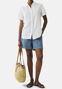 Camicia in lino bianca a maniche corte, pantaloni corti in denim blu, sandali neri e una borsa in paglia intrecciata con tracolla in pelle beige. Posizione eretta, sfondo neutro.