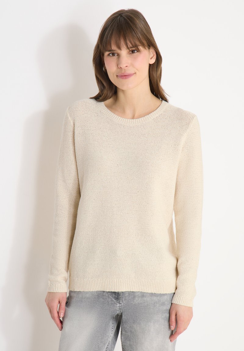 Femme aux cheveux bruns portant un pull en tricot couleur crème et un jean gris clair, debout devant un fond blanc.