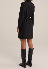 Svart skräddarsydd kappa med en åtsittande midja, långa ärmar och en minimalistisk design, i kombination med knähöga svarta läderstövlar. Slät textur.