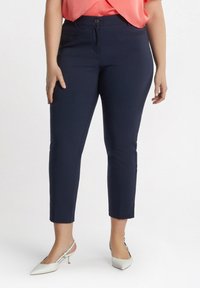 Marineblaue, knöchellange Hose mit glatter Textur, ausgestattet mit Vordertaschen und einem Knopfverschluss, kombiniert mit hellen Slingback-Heels.