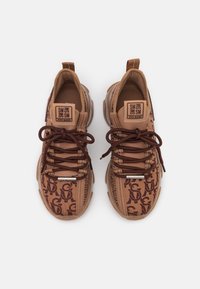 Paire de baskets marron Steve Madden avec des lacets marron foncé et un motif répétitif du logo SM sur les dessus en tissu.