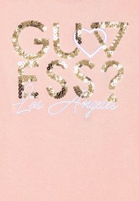 Chemise rose clair avec le texte en sequins dorés "GUESS?" et "Los Angeles" entouré d'un cœur en broderie blanche.
