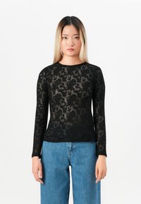 Ungdom med langt ombre-hår iført en gjennomsiktig svart blomsterdusk langermet topp og løse blå jeans, står mot en hvit bakgrunn.