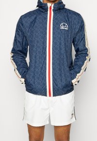Námorně modrá větrovka s geometrickým vzorem, červeným zipem a bílými detaily podél rukávů a lemu. Nastavitelná kapuce s detailem loga.