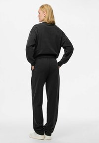 Kvinna med blont hår som bär en svart långärmad sweatshirt och matchande byxor, står med händerna i fickorna, vänd bort. Vita sneakers.