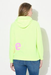 Neon groene hoodie met een roze letter "e" op de achterkant. Bevat een kangoeroe-pocket en ribgebreide manchetten. Gecombineerd met blauwe spijkerbroek.