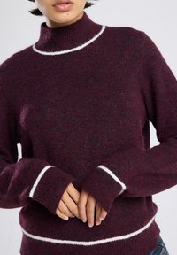 Persoon in een donkerpaars brei trui met witte afwerking op de kraag, manchetten en zoom, met handen zachtjes voor zich ineen clasped.