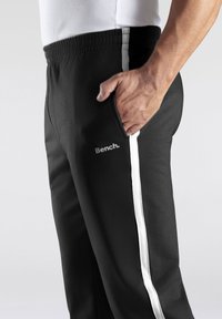 Schwarze Sporthose mit einem weißen Streifen an den Seiten, elastischem Bund und seitlicher Tasche. Mit dem "Bench."-Logo auf dem Oberschenkel.