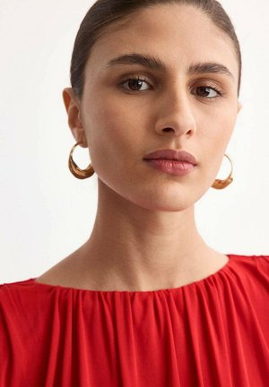 Primer plano de una persona con una blusa roja fruncida y un escote amplio. Pendientes de aro dorados realzan el look sobre un fondo neutro.