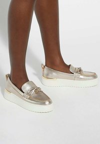 Metallic gold slip-on shoes with a textured finish, white sole, and decorative bow detail. Rounded shape and padded collar for comfort.