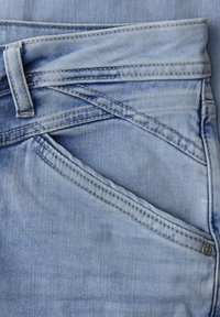 Poche de jean en denim bleu clair avec couture détaillée, passant de ceinture et rivet métallique sur le côté droit.