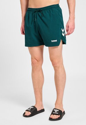 Mężczyzna noszący ciemnozielone sportowe spodenki z bocznymi rozcięciami i czarne klapki z białymi logo, stojący na jasnym tle.