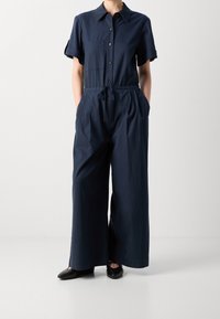 Granatowy kombinezon z lekkiego materiału, z krótkimi rękawami, zapięciem na guziki z przodu, regulowanym paskiem w talii oraz dwoma bocznymi kieszeniami.