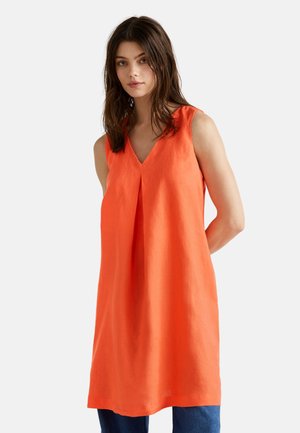 Ärmelloses orangenes Leinenkleid mit V-Ausschnitt und Falten-Detail an der Vorderseite, in einer lockeren, geraden Silhouette geschnitten, kombiniert mit blauen Jeans.