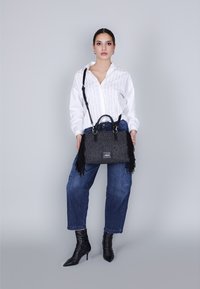 Borsa nera con frange realizzata in tessuto testurizzato, dotata di doppio manico e tracolla staccabile, abbinata a una camicia a righe bianche e jeans blu.