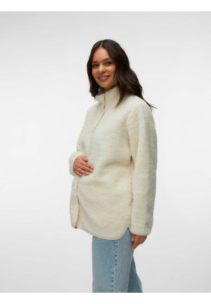 Donna con capelli lunghi e scuri che indossa una giacca in pile color crema e jeans azzurri, sorridente, che tiene la giacca vicino alla pancia.