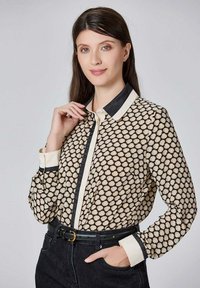 Blouse noire et crème avec un motif en coquillage, dotée d'un col noir contrastant et d'une patte de boutonnage. Manches longues avec des poignets crème.
