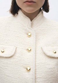 Chaqueta blanca texturizada con cuello mandarín, que presenta botones dorados y dos bolsillos frontales. La tela tiene un patrón en relieve.