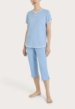 Donna che indossa una maglia a maniche corte celeste a pois con scollo a goccia e pantaloni corti abbinati, in piedi a piedi nudi su sfondo bianco.