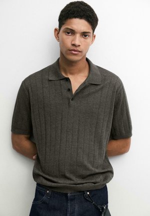 Jeune homme aux cheveux courts et bouclés, portant un polo côtelé à manches courtes de couleur vert olive foncé et un jean bleu foncé, debout contre un mur blanc.