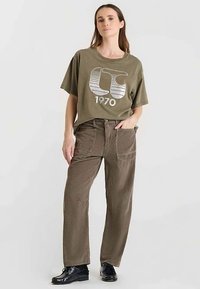 T-shirt vert olive avec un motif graphique à rayures blanches et le texte "1970", associé à un pantalon en velours côtelé marron avec poches et coupe décontractée.