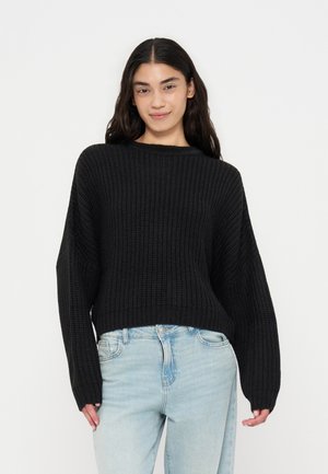 Kvinde med langt mørkt hår iført en sort chunky striktrøje og lyseblå jeans, står foran en ensfarvet lys baggrund.