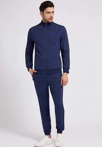 Navy blue athletic tracksuit with full zip, ribbed cuffs, and contrasting side stripes. Paired with white sneakers, model stands against a plain backdrop.