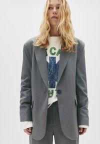 Blazer gri, croit personalizat dintr-un material texturat, cu închidere cu un singur buton. Purtați-l peste un hanorac grafic cu detalii imprimate verzi și albastre.