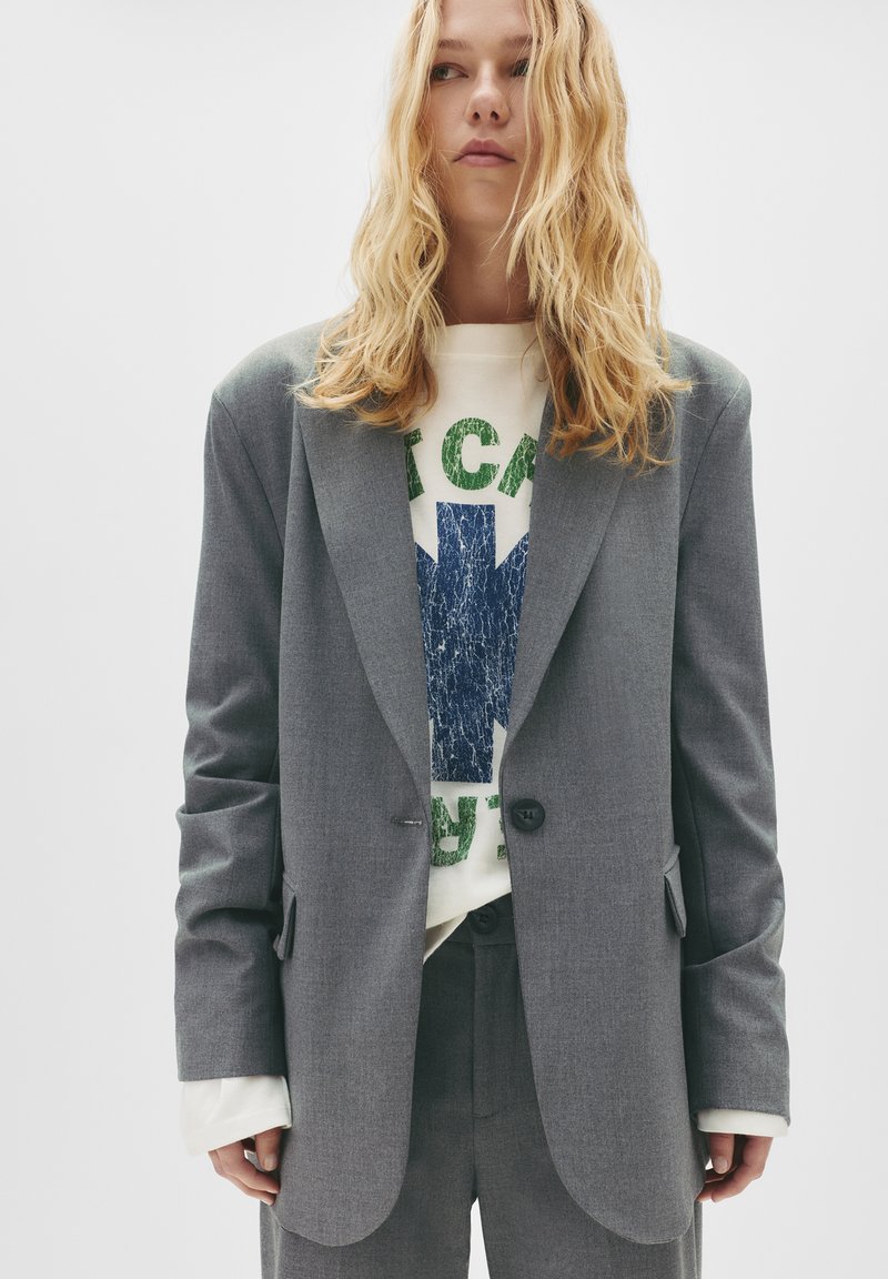 Blazer gri, croit personalizat dintr-un material texturat, cu închidere cu un singur buton. Purtați-l peste un hanorac grafic cu detalii imprimate verzi și albastre.