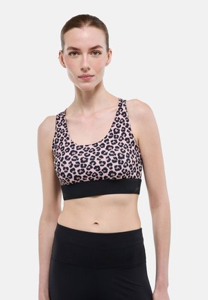 Femme portant un soutien-gorge de sport imprimé léopard rose et noir et un legging noir taille haute, debout devant un fond blanc uni.