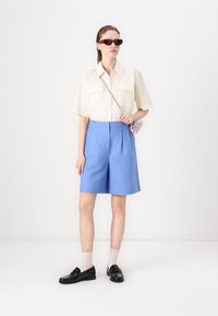 Camicia corta beige chiaro con maniche corte, pantaloni tailored blu con pieghe frontali, mocassini neri e handbag rosa chiaro, completato da occhiali da sole rossi.