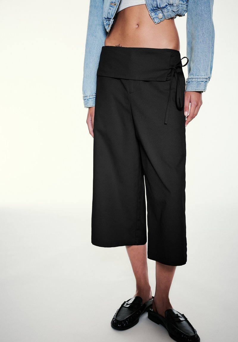 Femme portant un pantalon noir large et court avec une taille nouée, des mocassins noirs et une veste courte en jean, laissant voir son ventre.